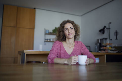 Mittlere erwachsene Frau trinkt Kaffee, während sie zu Hause am Tisch sitzt - RBF07968