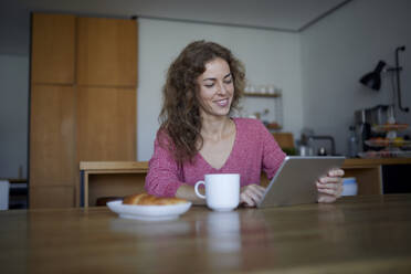 Frau mit Essen und Kaffee, die ein digitales Tablet benutzt, während sie zu Hause sitzt - RBF07969