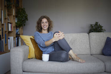 Smiling woman drinking coffee while sitting on sofa at home - RBF07973