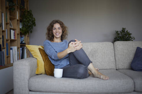 Lächelnde Frau trinkt Kaffee, während sie zu Hause auf dem Sofa sitzt - RBF07973