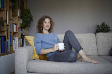 Frau hält Kaffeetasse, während sie zu Hause auf dem Sofa sitzt - RBF07974