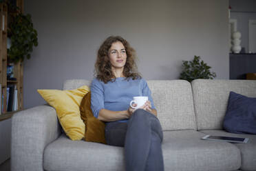 Lächelnde Frau mit Kaffeetasse, die wegschaut, während sie zu Hause auf dem Sofa sitzt - RBF07975