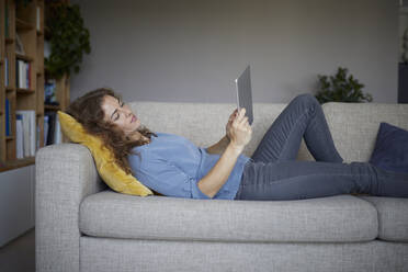 Frau benutzt digitales Tablet, während sie zu Hause auf dem Sofa liegt - RBF07978