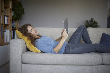Woman using digital tablet while lying on sofa at home - RBF07978