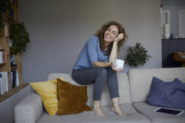 Smiling woman drinking coffee while sitting on top of sofa at home - RBF07979