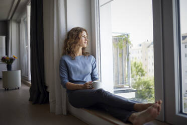 Woman holding coffee cup while sitting on window sill at home - RBF07983