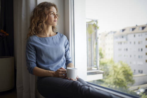 Frau mit Kaffeetasse, die durch das Fenster schaut, während sie auf der Fensterbank zu Hause sitzt - RBF07984