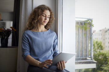 Frau benutzt digitales Tablet, während sie zu Hause auf der Fensterbank sitzt - RBF07985