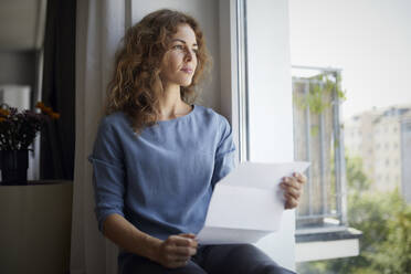 Woman holding paper while sitting on window sill at home - RBF07986