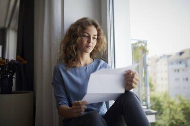 Frau liest Zeitung, während sie zu Hause auf der Fensterbank sitzt - RBF07987