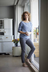 Smiling woman holding coffee cup while leaning on door by balcony at home - RBF07989