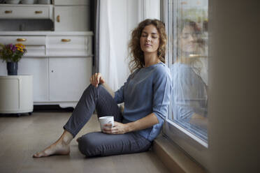 Mid adult woman with coffee cup sitting while leaning on glass door at home - RBF07990