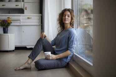 Mittlere erwachsene Frau mit Kaffeetasse, die sich zu Hause an eine Glastür lehnt - RBF07990