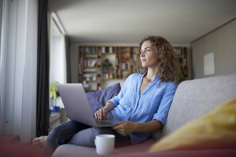 Frau benutzt Laptop, während sie zu Hause auf dem Sofa sitzt, lizenzfreies Stockfoto