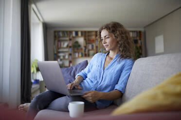 Mid adult woman using laptop while sitting on sofa at home - RBF07997