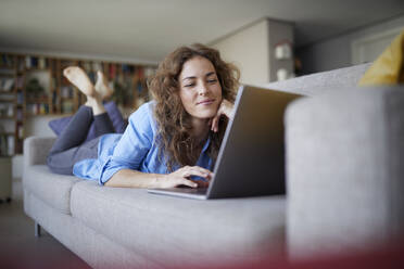 Lächelnde Frau, die einen Laptop benutzt, während sie zu Hause auf dem Sofa liegt - RBF07999