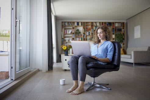 Mittlere erwachsene Frau arbeitet am Laptop, während sie zu Hause auf einem Stuhl sitzt - RBF08001