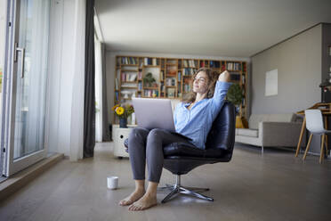Smiling woman with hands behind head using laptop while sitting on chair at home - RBF08002