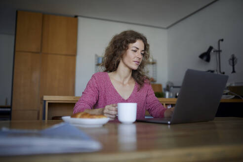 Frau frühstückt, während sie zu Hause am Laptop arbeitet - RBF08004