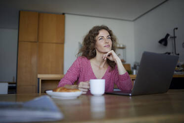 Nachdenkliche Frau mit Laptop und Kaffeetasse, während sie zu Hause sitzt - RBF08005