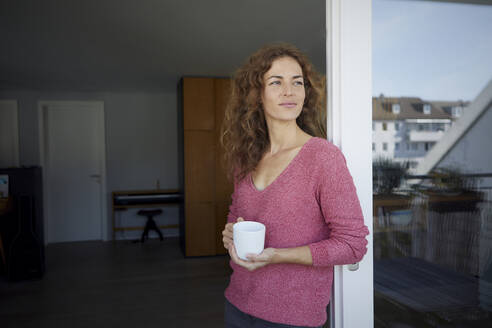 Schöne Frau hält Kaffeetasse und lehnt sich an die Tür zu Hause - RBF08013