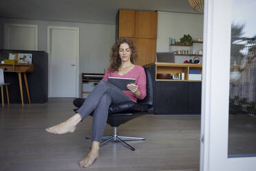 Mittlere erwachsene Frau, die ein digitales Tablet benutzt, während sie zu Hause auf einem Stuhl sitzt - RBF08016