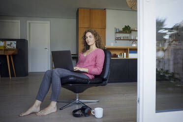 Mittlere erwachsene Frau arbeitet am Laptop, während sie zu Hause auf einem Stuhl sitzt - RBF08018