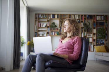 Mittlere erwachsene Frau, die einen Laptop benutzt, während sie zu Hause auf einem Stuhl sitzt - RBF08023