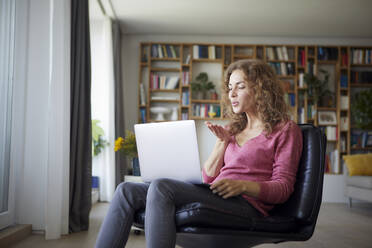 Frau bläst Kuss zu Videoanruf auf Laptop, während sie zu Hause sitzt - RBF08024