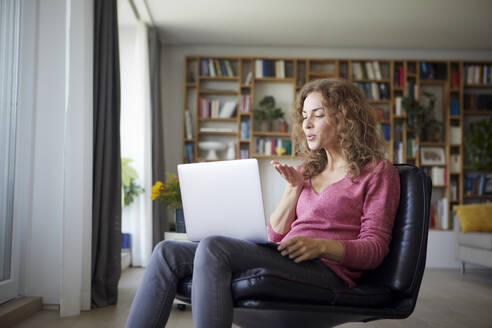 Frau bläst Kuss zu Videoanruf auf Laptop, während sie zu Hause sitzt - RBF08024
