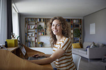 Lächelnde Frau arbeitet am Laptop, während sie zu Hause am Schreibtisch sitzt - RBF08028