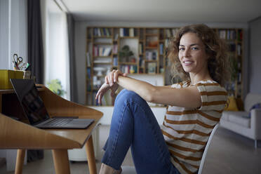 Woman smiling while sitting by desk on chair at home - RBF08032