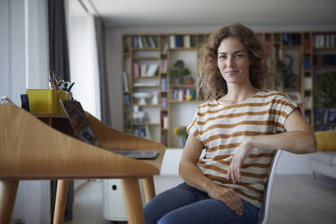 Lächelnde Frau im mittleren Erwachsenenalter, die zu Hause auf einem Stuhl am Schreibtisch sitzt - RBF08033