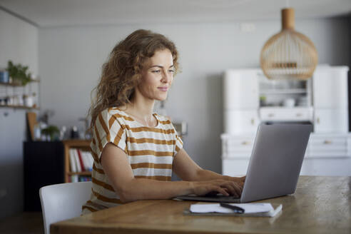 Mittlere erwachsene Frau arbeitet am Laptop, während sie zu Hause am Tisch sitzt - RBF08034