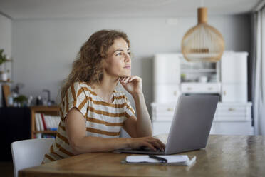 Nachdenkliche Frau mit Kopf in den Händen, die einen Laptop benutzt, während sie zu Hause am Tisch sitzt - RBF08035