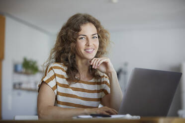 Lächelnde Frau im mittleren Erwachsenenalter, die einen Laptop benutzt, während sie zu Hause sitzt - RBF08036