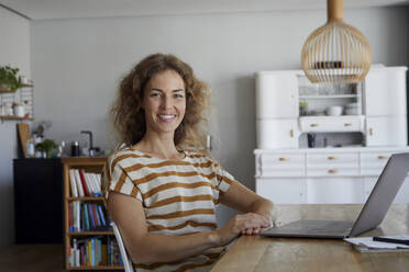 Smiling woman with laptop sitting by table at home - RBF08037