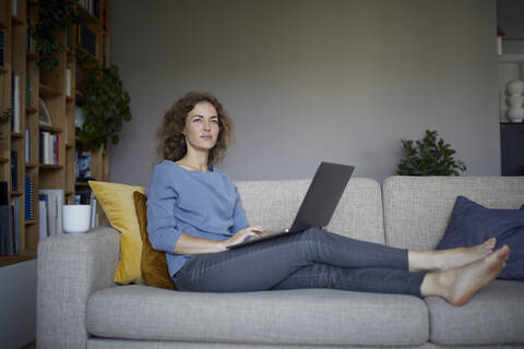 Frau schaut weg, während sie auf dem Sofa zu Hause sitzt und einen Laptop benutzt, lizenzfreies Stockfoto