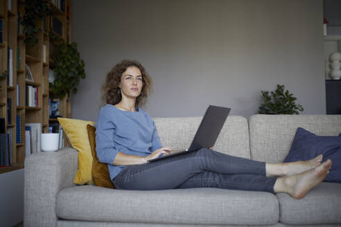 Frau schaut weg, während sie auf dem Sofa zu Hause sitzt und einen Laptop benutzt - RBF08044