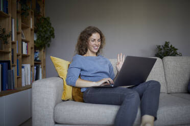 Mittlere erwachsene Frau winkt mit der Hand zum Videoanruf auf dem Laptop, während sie zu Hause sitzt - RBF08046