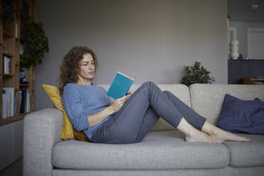 Frau liest ein Buch, während sie zu Hause auf dem Sofa sitzt - RBF08047