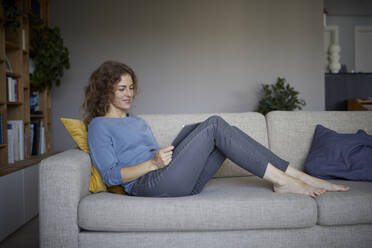 Lächelnde Frau, die ein digitales Tablet benutzt, während sie zu Hause auf dem Sofa sitzt - RBF08048