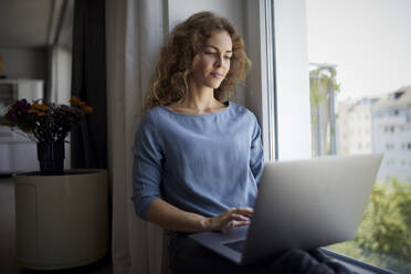 Frau benutzt Laptop, während sie zu Hause auf der Fensterbank sitzt - RBF08049