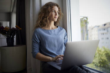 Mid adult woman using laptop while sitting on window sill at home - RBF08050