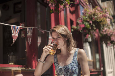 Woman drinking beer while sitting outside pub during sunny day - AJOF00164