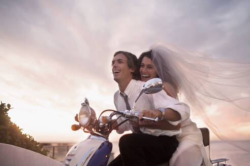 Cheerful newlywed couple in wedding dress riding motor scooter during sunset - AJOF00173