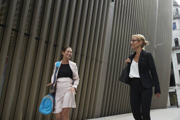 Smiling female colleagues talking while walking by office building in downtown - PMF01288