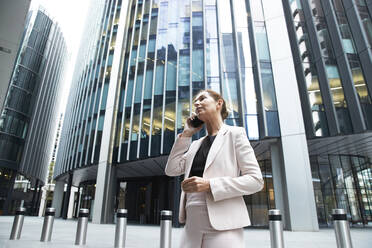 Businesswoman talking on mobile phone while standing against modern office skyscraper in financial district - PMF01297