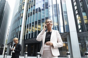 Female professional talking on mobile phone while standing near businesswoman against modern office building - PMF01298