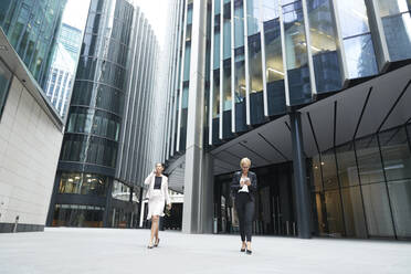 Female entrepreneurs walking with smart phones against office building at downtown district in city - PMF01300